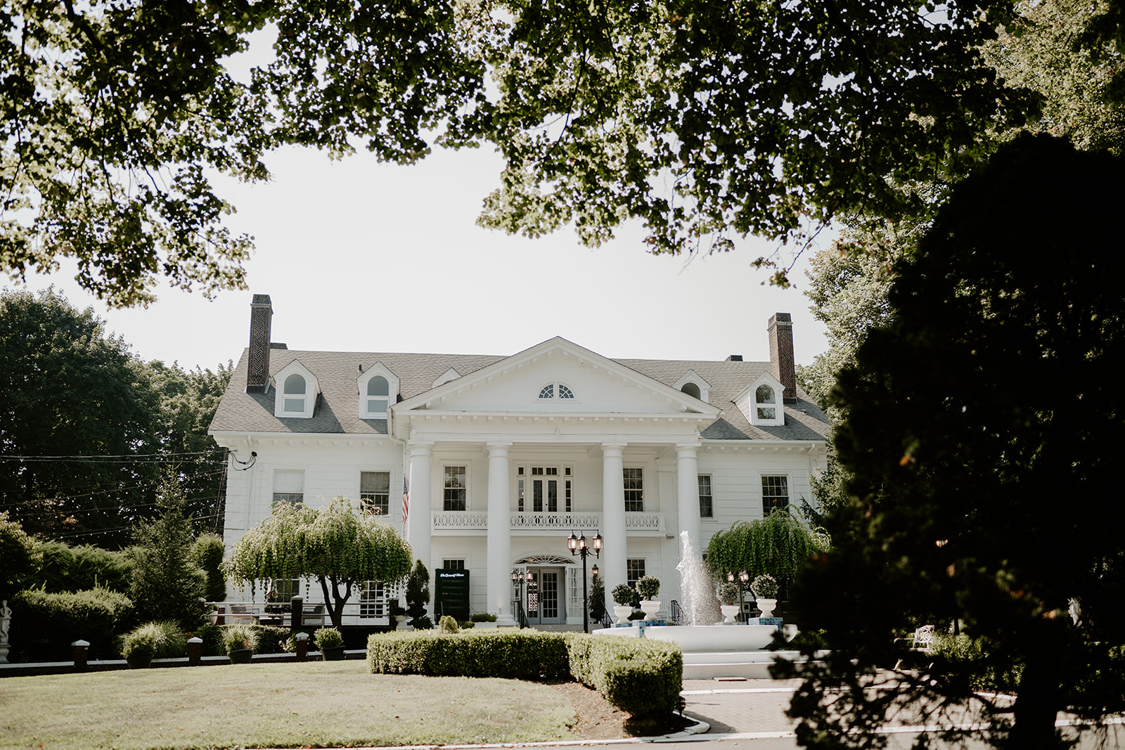 Wide landscape view of the grand white facade of The Briarcliff Manor, framed by lush green trees.